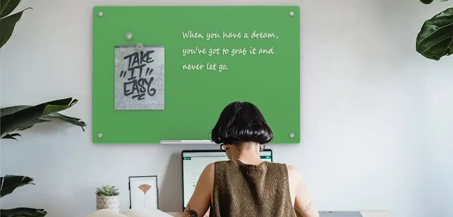 In a room with green plants and white walls, there's a green glass whiteboard hanging up. And there's a girl really getting down to work.