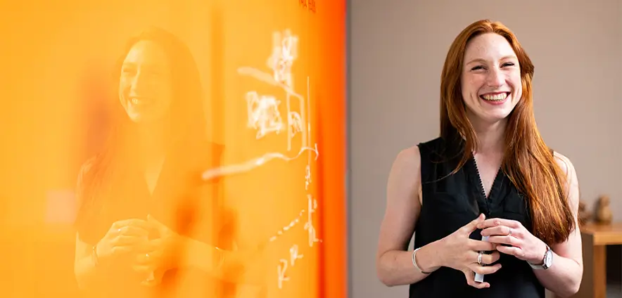 A women is standing in front of an orange glass whiteboard, happily discussing the words written on the whiteboard with someone.
